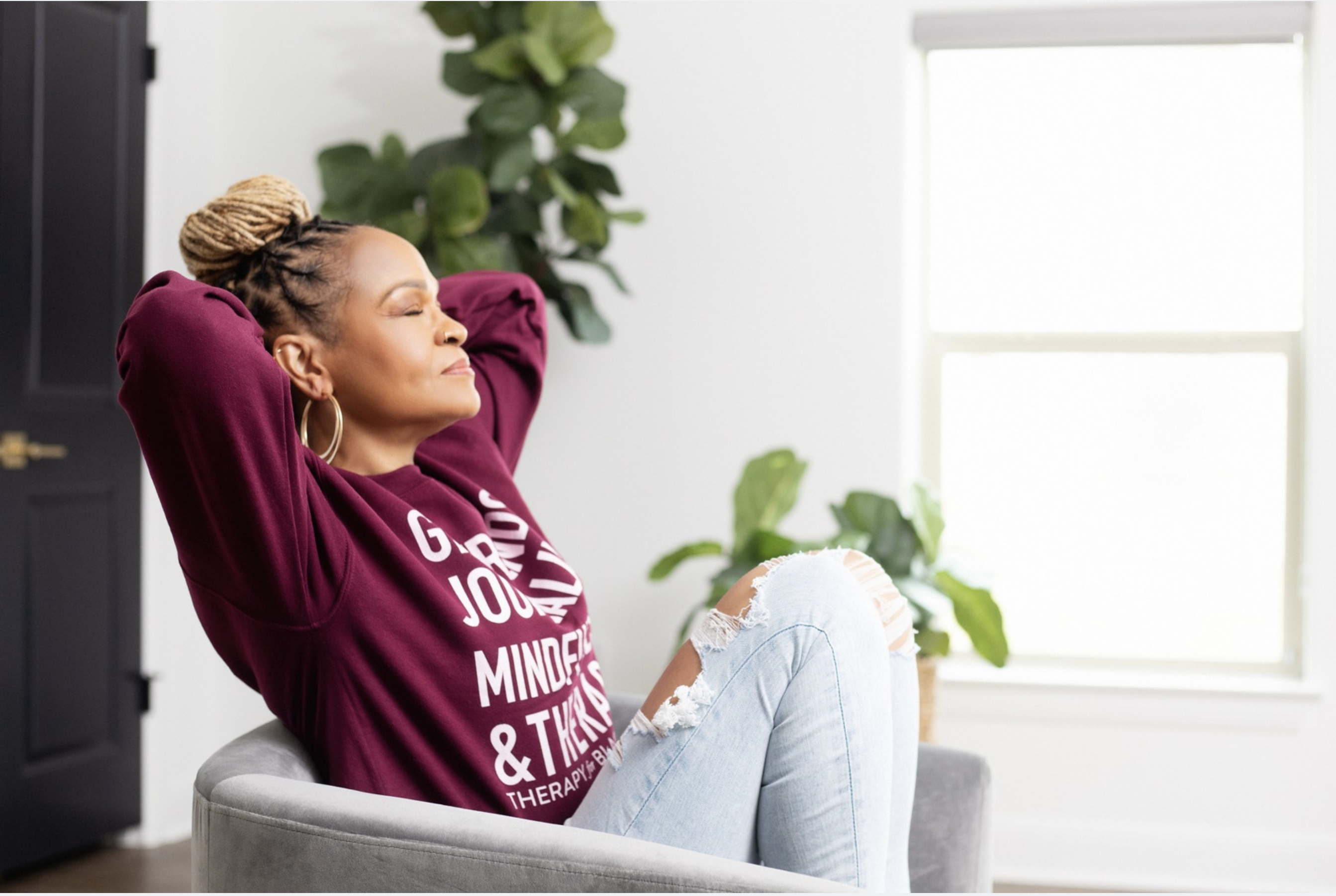 A woman with braided hair in a bun sits relaxed in a grey chair, wearing a burgundy sweatshirt and ripped jeans, with her eyes closed and hands behind her head, symbolizing a moment of peace and reflection in the journey of rewriting her internal script.
